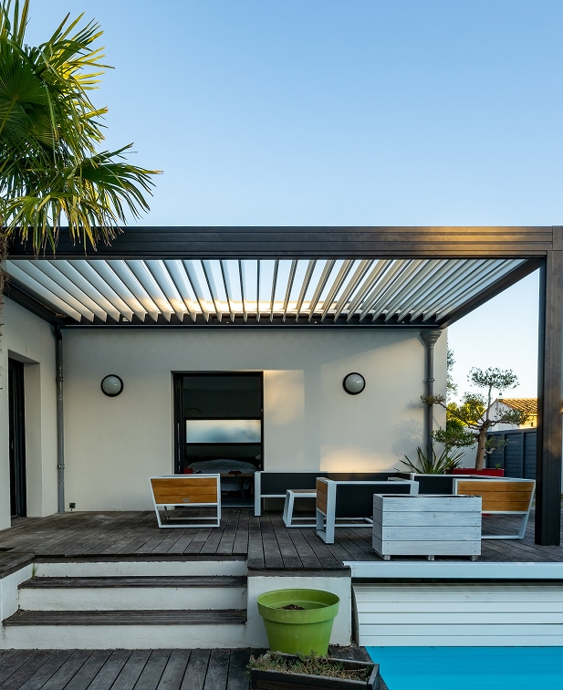 Pergola bioclimatique noire au-dessus d'une terrasse en bois. Mobilier moderne blanc/bois près de la piscine sous un ciel bleu clair.