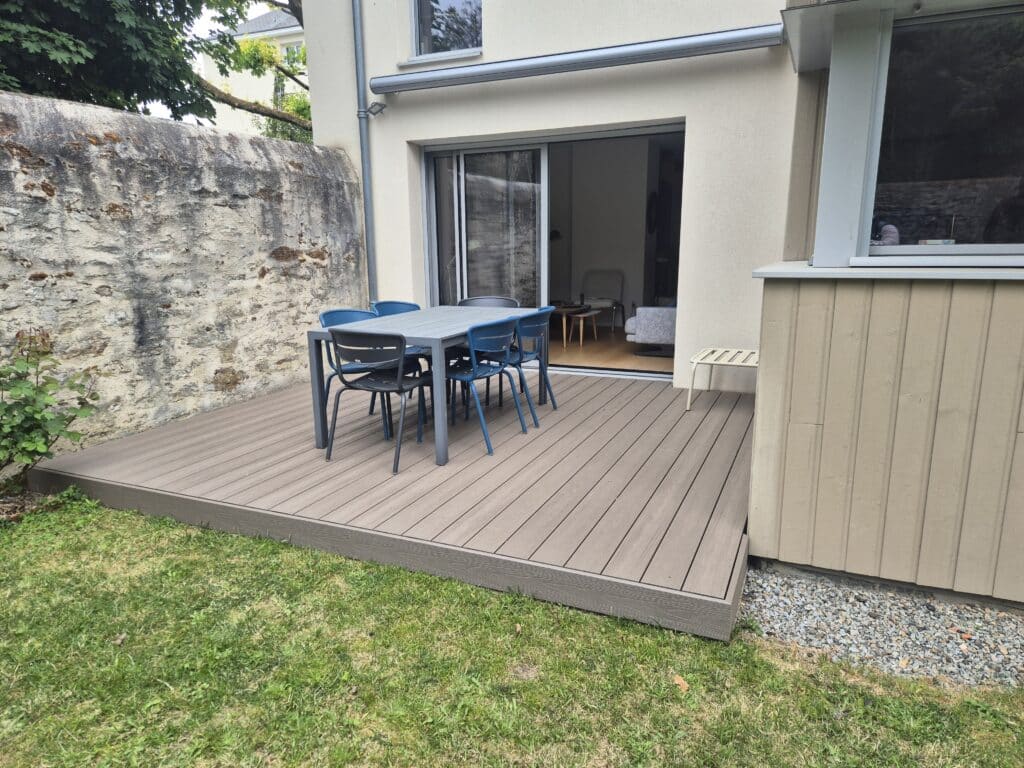 Terrasse en bois composite avec salon de jardin devant une maison moderne et un vieux mur en pierre.