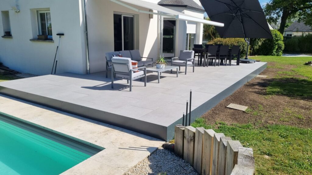 Terrasse moderne en dalles grises avec mobilier de jardin, parasol noir et piscine bordant une villa blanche.